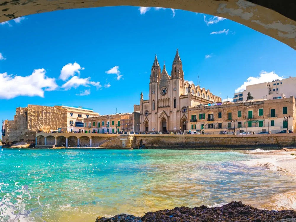 Balluta Bay in Malta, a church and terraced buildings above turquoise water, framed by a stone arch