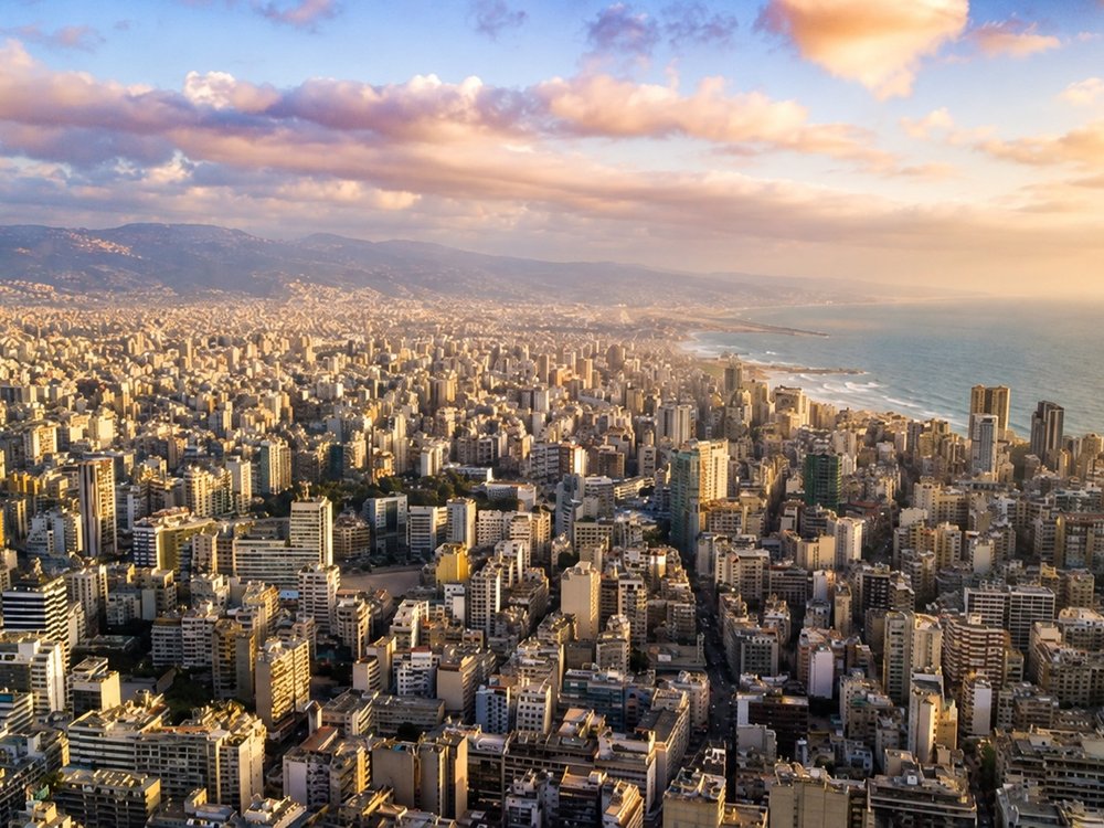 Aerial view of a Middle Eastern coastal city at dusk, dense apartment towers running along the shoreline
